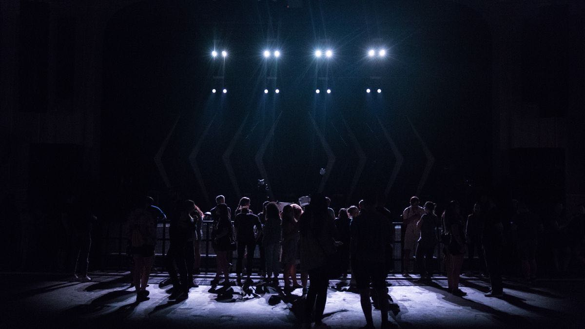 A crowd standing in near-darkness in front of a stage while four bright lights point over their heads