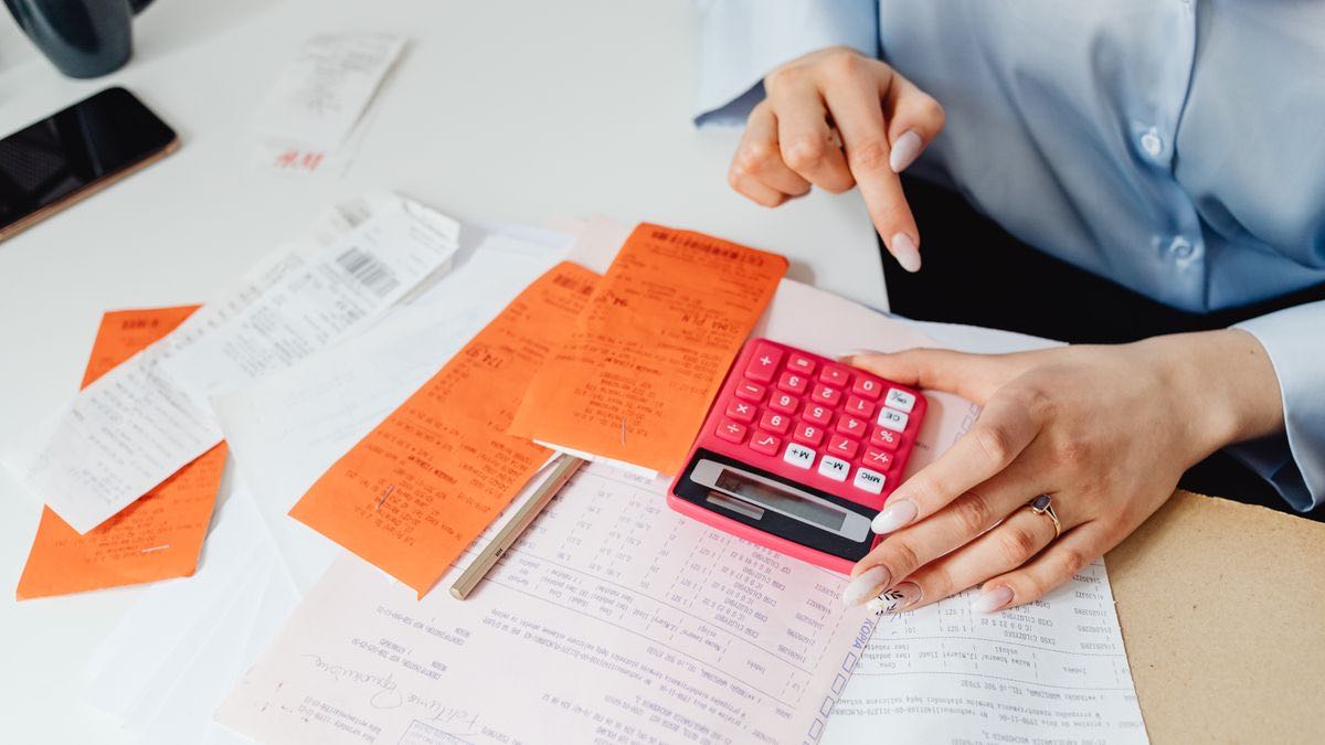 A woman at a white desk working through a pile of orange and white receipts with a pink calculator