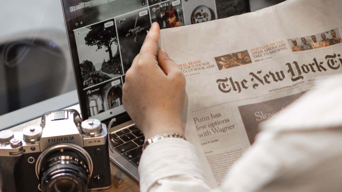A hand holding a newspaper beside a laptop and camera, illustrating news data feeding a monitoring tool