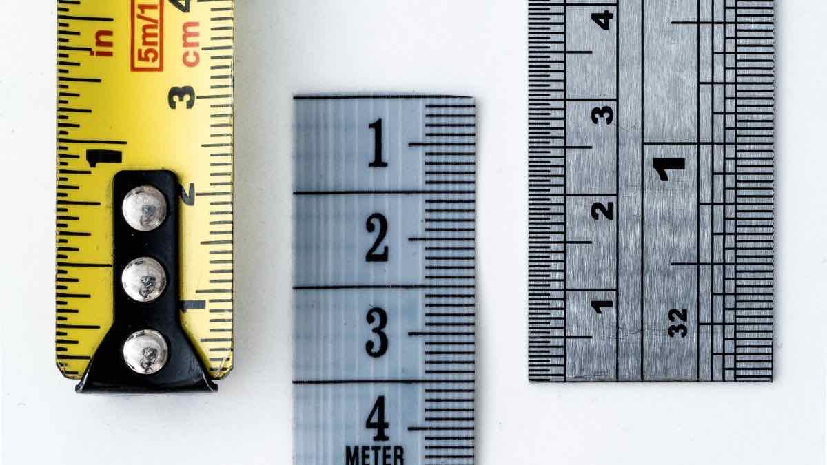 A yellow tape measure, a plastic ruler and a steel rule lying side by side on a white surface, each marked with inch and centimetre scales