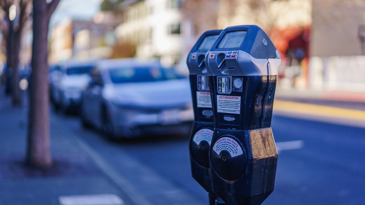 A pair of coin-and-card parking meters on a sunlit street, parked cars blurred behind them