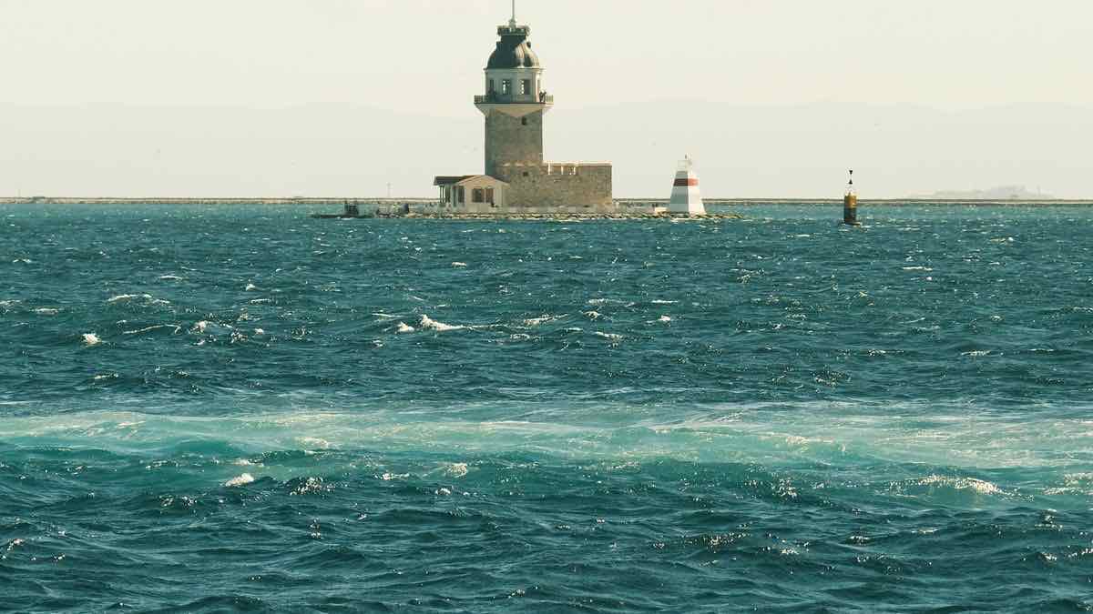 A stone tower lighthouse on a breakwater across choppy turquoise water, two small navigation markers beside it