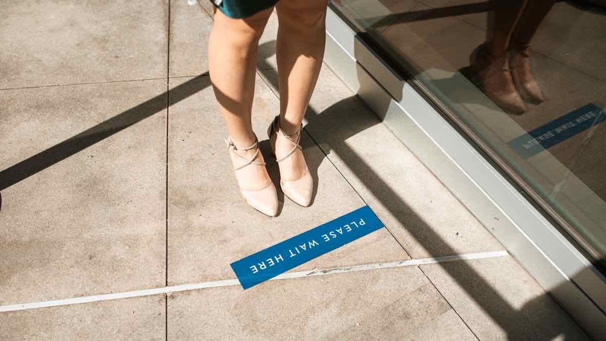 Person standing behind a blue 'please wait here' floor marker outside a glass door