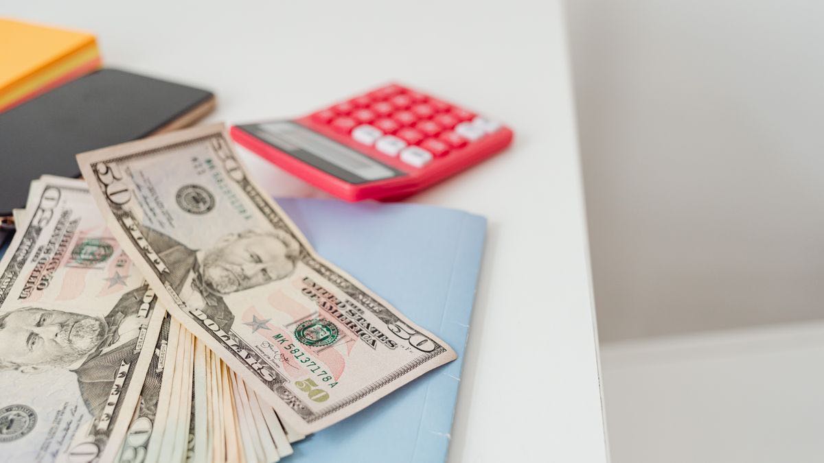 Several US dollar bills fanned out on a white desk beside a red desk calculator