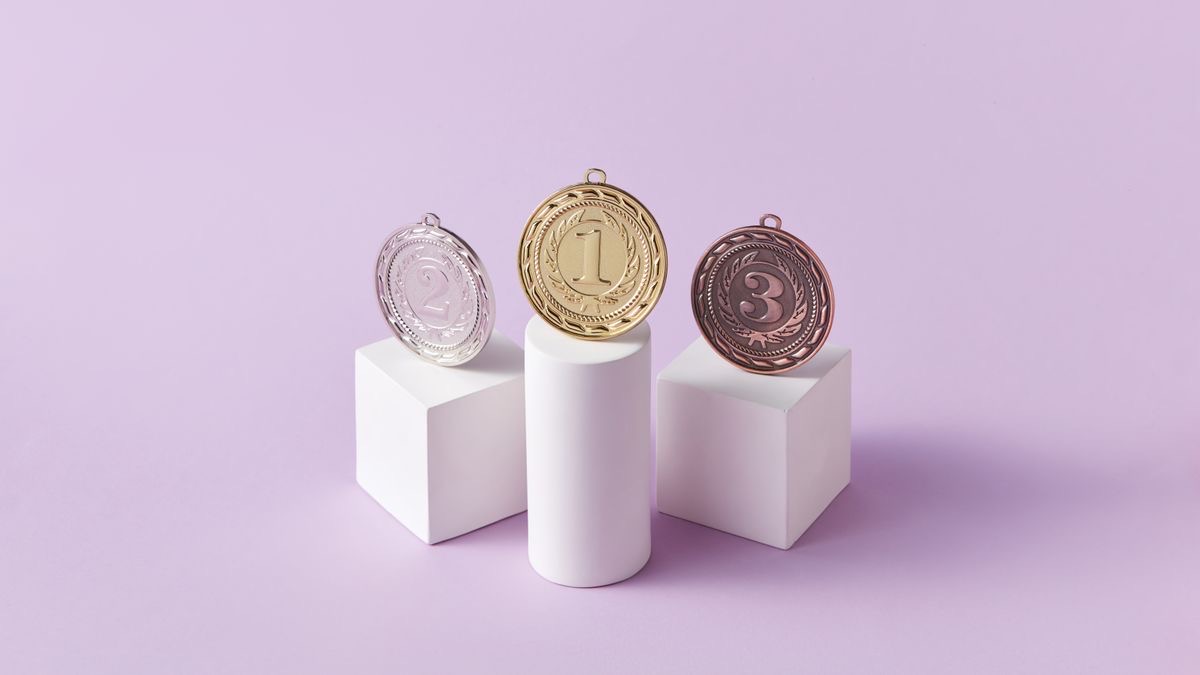 A gold, silver and bronze medal, each standing on its own white pedestal against a pale purple background