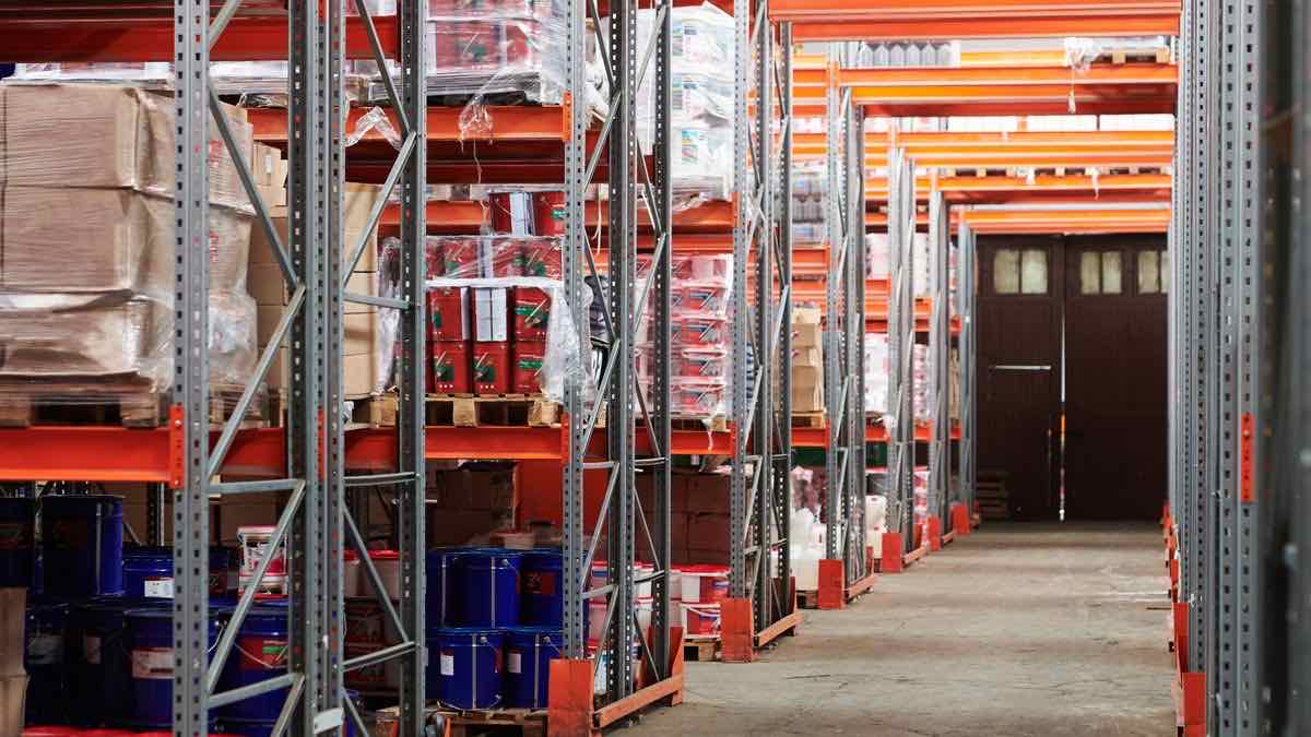 A long warehouse aisle between tall shelving racks stacked with cardboard boxes and pails