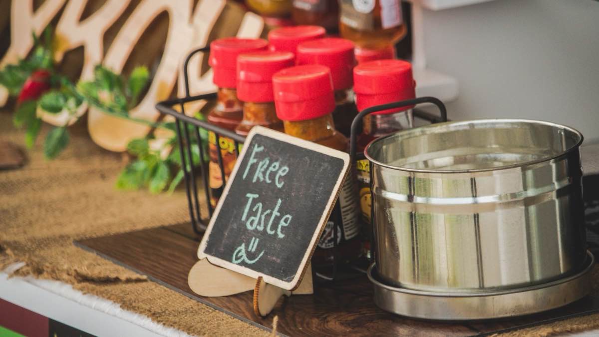 A small chalkboard sign reading Free Taste beside a basket of sauce bottles on a market table