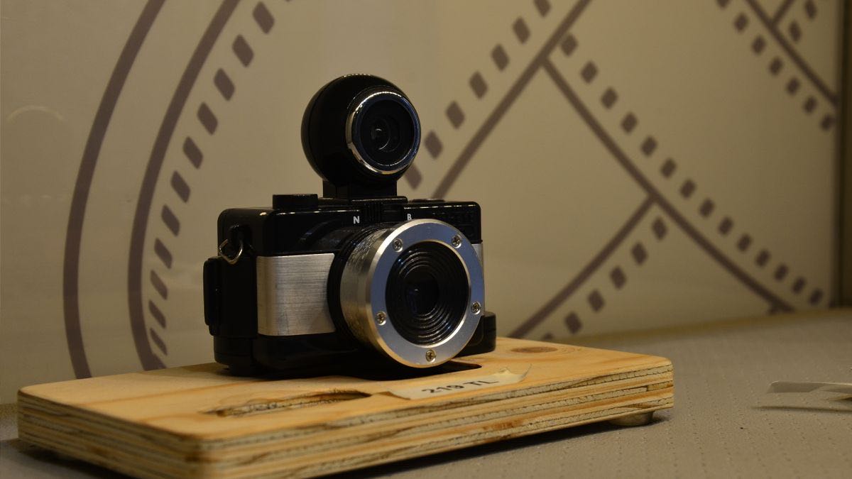 A black compact film camera with a stacked lens and a ring finder, resting on a small wooden board against a film-strip pattern