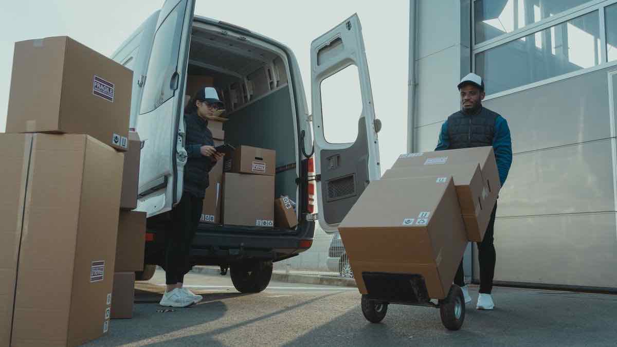 Two couriers unloading cardboard boxes from an open white van, one pushing a loaded hand truck
