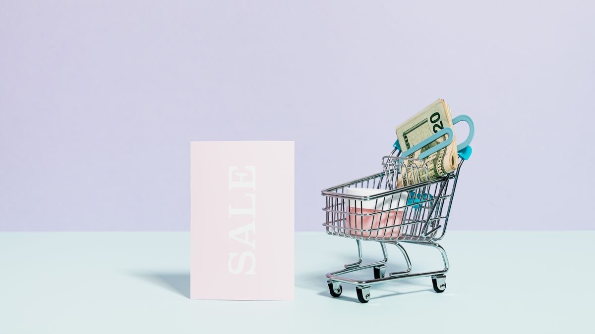 A miniature metal shopping cart holding a roll of banknotes beside a pale pink card that reads SALE