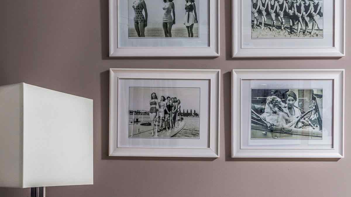 Four framed black-and-white photographs of people at a beach and in a car hung on a mauve wall beside a lamp