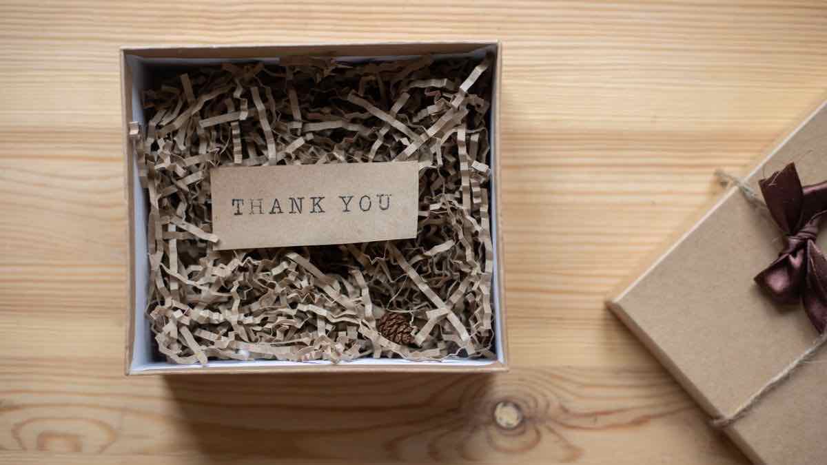An open cardboard gift box holding a Thank You card in shredded paper, beside a wrapped parcel on a wooden table