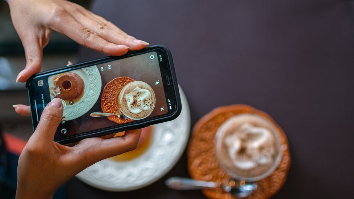 Two hands holding a phone above a cafe table, framing a dessert and a latte in the camera