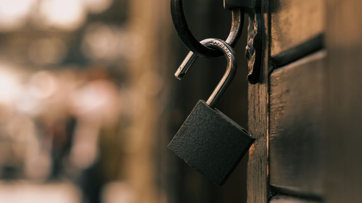 An unlocked padlock hanging open from a latch on a dark wooden door, standing for access that is free but gated