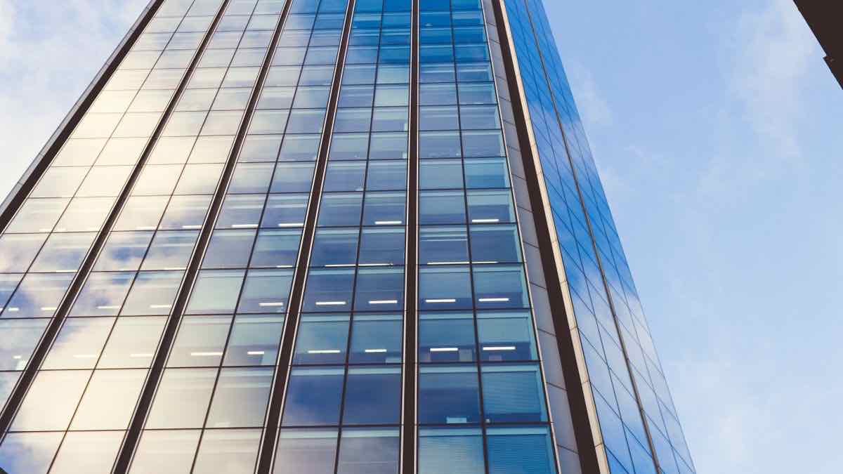 Low-angle view of a glass office tower with lit windows against a pale blue sky