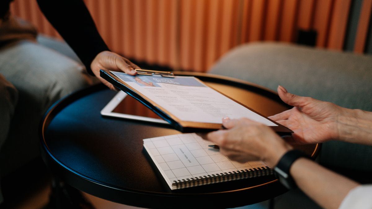A hand passing a clipboard with a printed resume across a small round table, with a notebook and tablet nearby