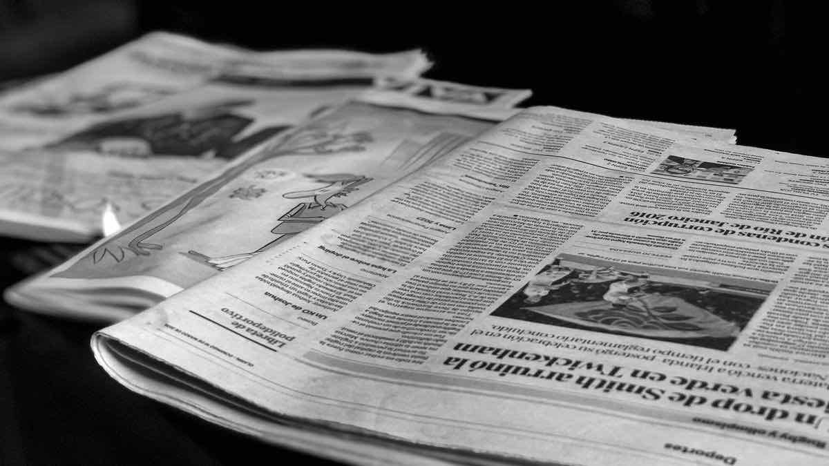 Black-and-white photo of several newspapers laid open on a table, a printed article visible in the foreground