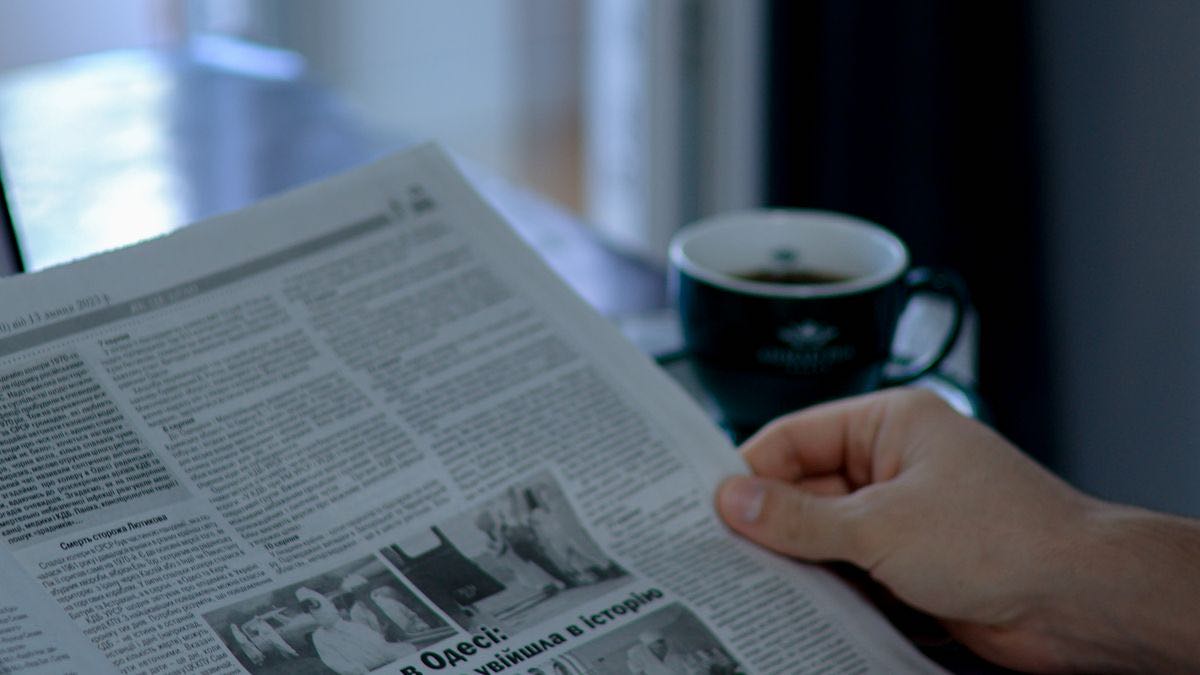 A hand holding an open newspaper beside a dark coffee cup
