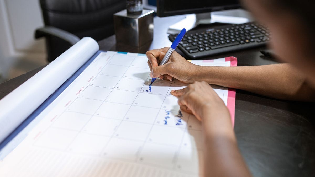 A hand writing with a blue pen on a monthly desk calendar, with a keyboard and monitor behind it