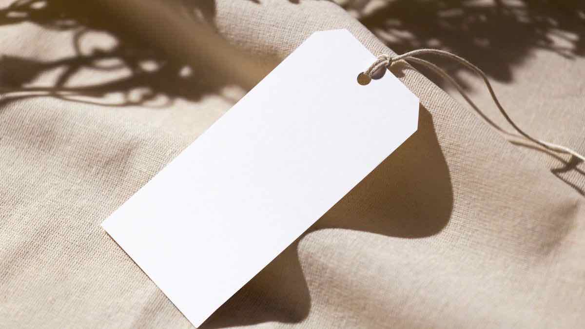A blank white paper price tag on a string, resting on beige fabric in dappled light