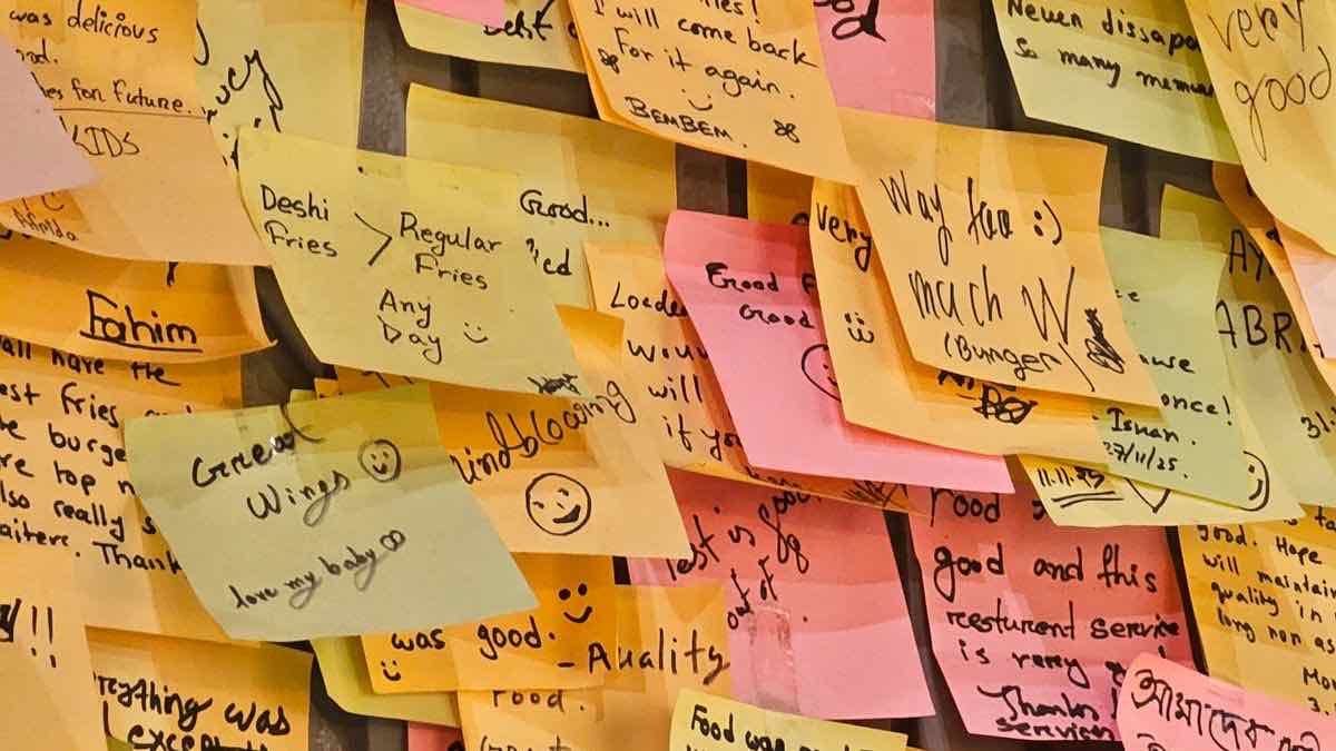 A wall crowded with overlapping handwritten sticky notes in yellow, orange and pink, each from a different person