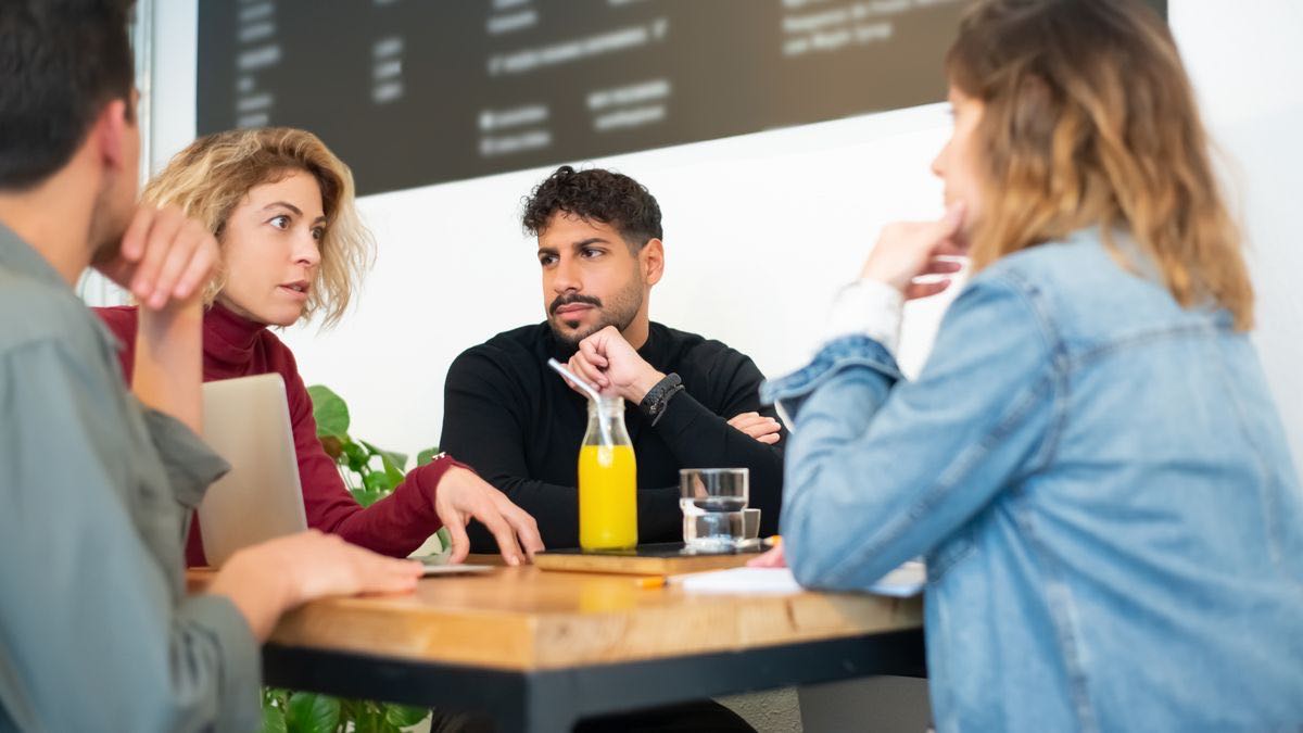 Four people seated around a wooden cafe table in conversation, two of them resting their chins on their hands