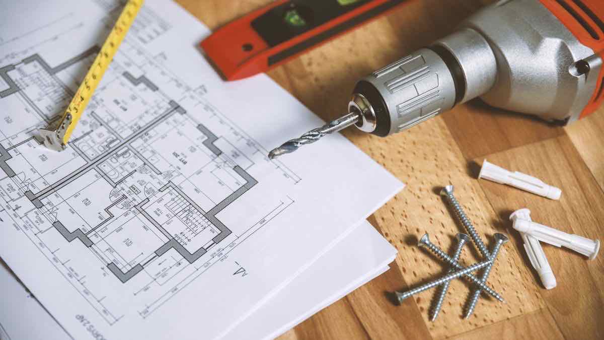A house floor plan on a wooden table with a yellow tape measure, a spirit level, a drill and a few screws