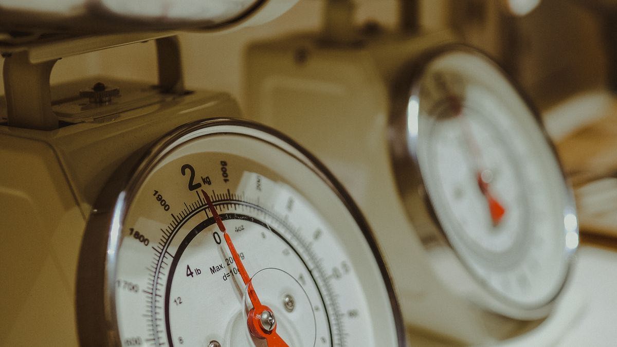 Close-up of the dial of a cream kitchen scale with a red needle, a second scale blurred behind it