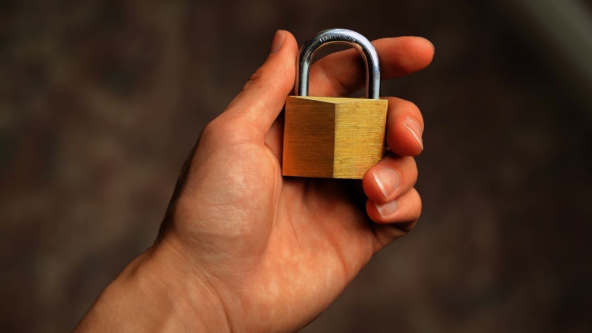 A hand holding a small brass padlock against a dark blurred background, standing for access that is gated rather than open