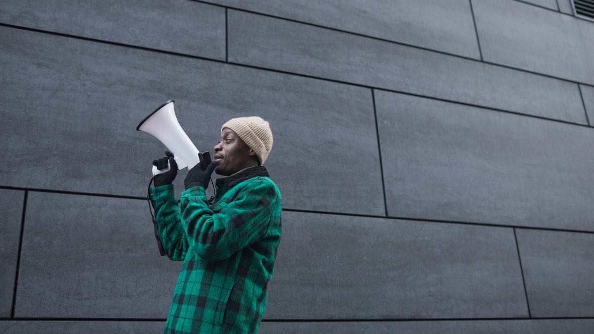 A man in a green plaid jacket and beanie speaking through a white megaphone in front of a grey panelled wall