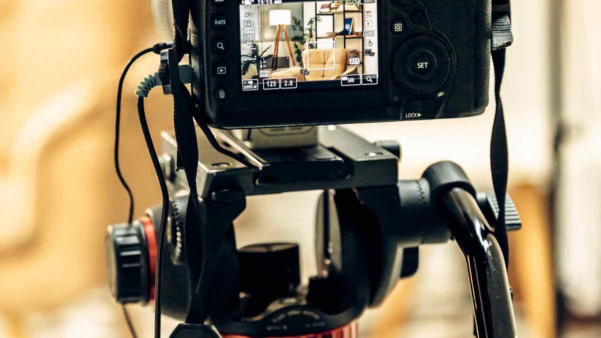 A video camera on a tripod head with its monitor lit, showing a living room scene being framed
