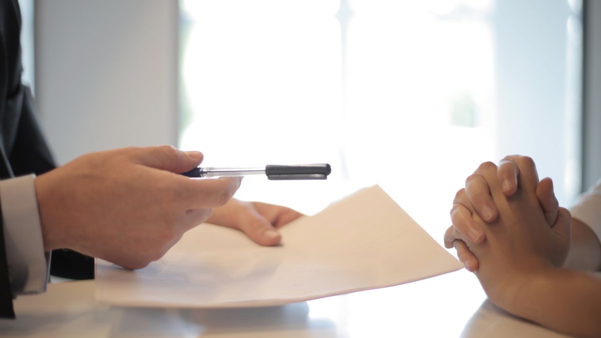 A hiring manager handing a pen and document across a desk in an interview — the end of the sourcing funnel