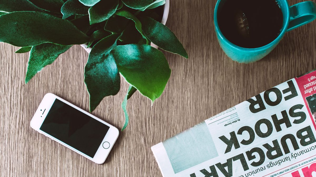 A phone beside a newspaper with a headline, representing a Google News feed of fresh articles